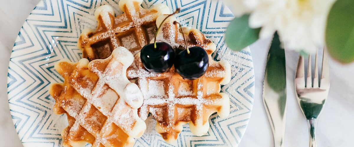 3 waffles on saucer with 2 cherry fruits beside fork and bread knife